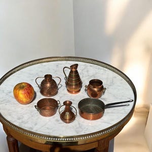 May include: A selection of miniature copper kitchen utensils, such as a saucepan, jugs, and bowls, displayed on a marble-topped table. A red and yellow apple is also present. The items are on a round table with a brass edge.