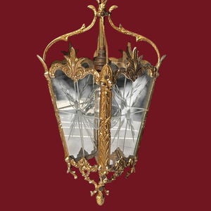 Könnte beinhalten: Verzierte, goldfarbene Hängelaterne mit klaren Glasscheiben. Das Glas ist mit einem blattartigen Muster geätzt. Die Laterne hat einen dekorativen Rahmen im Filigranstil und wird an einer Kette aufgehängt. Der Hintergrund ist einfarbig weinrot.
