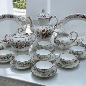 Könnte beinhalten: Ein weißes Porzellan-Teeservice mit zartem Blumenmuster aus roten Mohnblumen und blauen Wildblumen sowie goldfarbenen Rändern, bestehend aus einer Teekanne, einer Zuckerdose mit Deckel, einem Milchkännchen, mehreren Tassen mit Untertassen und zwei ovalen Platten.