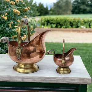 Può includere: Un set di due zuccheriere in rame e ottone con manici decorativi. La ciotola più grande presenta un design a testa di leone e la ciotola più piccola ha un design floreale.