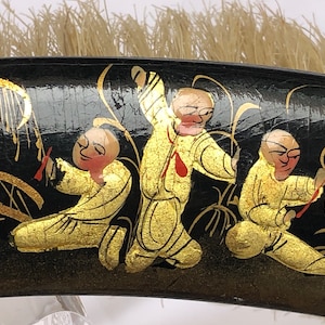 Peut inclure: Un peigne à cheveux en laque noire avec des figures peintes en or et en rouge. Les figures sont habillées de robes jaunes et sont représentées de manière stylisée. Le peigne a une brosse au dos.