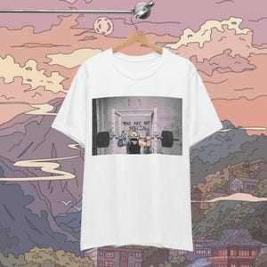 May include: White t-shirt with a graphic of a cartoon character lifting weights in a gym. The text "You are not special" is written on the wall behind the character.