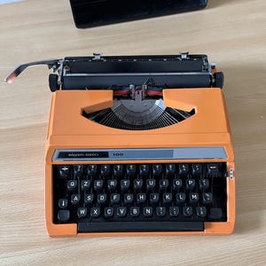 Puede incluir: Una máquina de escribir vintage Silver-Reed 100 de color naranja con un teclado negro y una placa de identificación plateada. La máquina de escribir tiene una palanca de retorno de carro y un rodillo de papel.