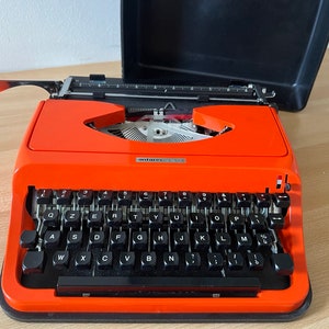 May include: An orange vintage Olivetti Lettera 32 typewriter with a black keyboard and a black carrying case. The typewriter has a silver return lever and a silver carriage return key.