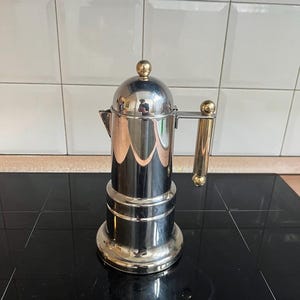 May include: A stainless steel stovetop espresso maker with a gold-colored handle and top knob. The coffee pot is cylindrical with a rounded top and a spout. The pot is sitting on a black stovetop.
