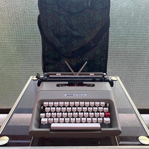 May include: A vintage Olivetti Lettera 35 typewriter in light grey, with white keys and black lettering, plus a red return key. It sits on a glass table, with a black carrying case behind it. The typewriter is a classic writing tool.