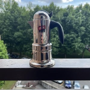 May include: A stainless steel stovetop espresso maker with a black handle and spout. The coffee pot has the text "VIGANO" on a red and white label. The espresso maker is sitting on a wooden railing with a green tree background.