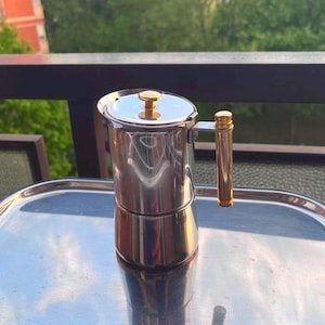 May include: A stainless steel stovetop espresso maker with a gold-colored handle and lid knob. The coffee pot is sitting on a silver tray, reflecting the sunlight. The background features a balcony railing and green trees.