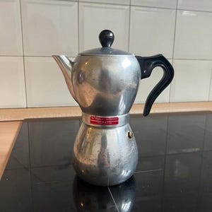 May include: A silver stovetop espresso maker with a black handle and lid. The coffee pot has a classic design, with a round base and a spout for pouring. The handle is curved for easy gripping. The pot is sitting on a black surface.
