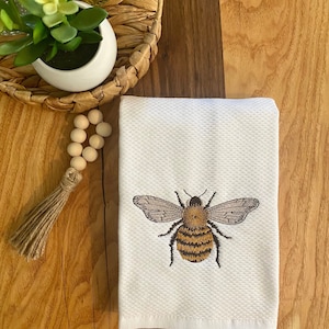May include: White hand towel with a textured pattern, featuring an embroidered bee design. The bee has brown and yellow stripes, with detailed wings. The towel is folded and placed on a wooden surface.