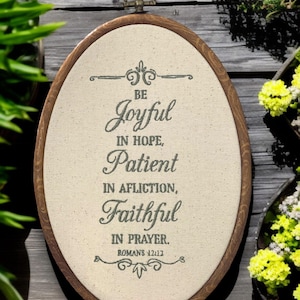 May include: An oval wooden framed artwork with the text "BE Joyful in Hope, Patient in Affliction, Faithful in Prayer. Romans 12:12" printed on a beige fabric. The frame has a metal loop for hanging.