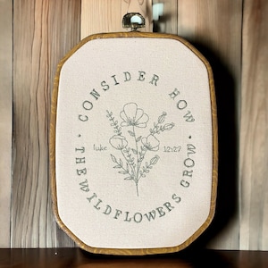 May include: Embroidered hoop art with a beige background and a brown wooden frame. The embroidery features a green floral design with the text "Consider how the wildflowers grow. Luke 12:27"