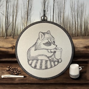 May include: Embroidered hoop art featuring a black and white raccoon holding a coffee cup. The raccoon is sitting on a beige background.