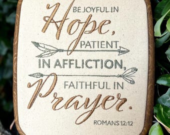 Könnte beinhalten: Ein gerahmtes Stickbild mit beigefarbenem Hintergrund und einem braunen Holzrahmen. Die Stickerei zeigt den Text "BE JOYFUL IN Hope. PATIENT IN AFFLICTION, FAITHFUL IN Prayer. ROMANS 12:12" in einer kursivschrift. Der Text ist in braunem Garn gestickt und die Pfeile in grünem Garn.