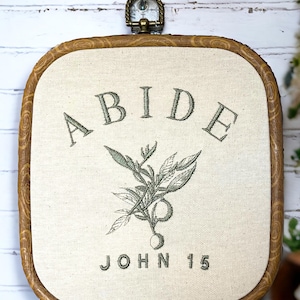 May include: A framed embroidery with a wooden frame and a white background. The embroidery features a green plant with the word "ABIDE" above it and "JOHN 15" below it.