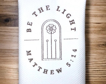May include: White kitchen towel with brown embroidery that reads "Be the Light Matthew 5:14". The embroidery also includes a stylized image of a window with a flower.