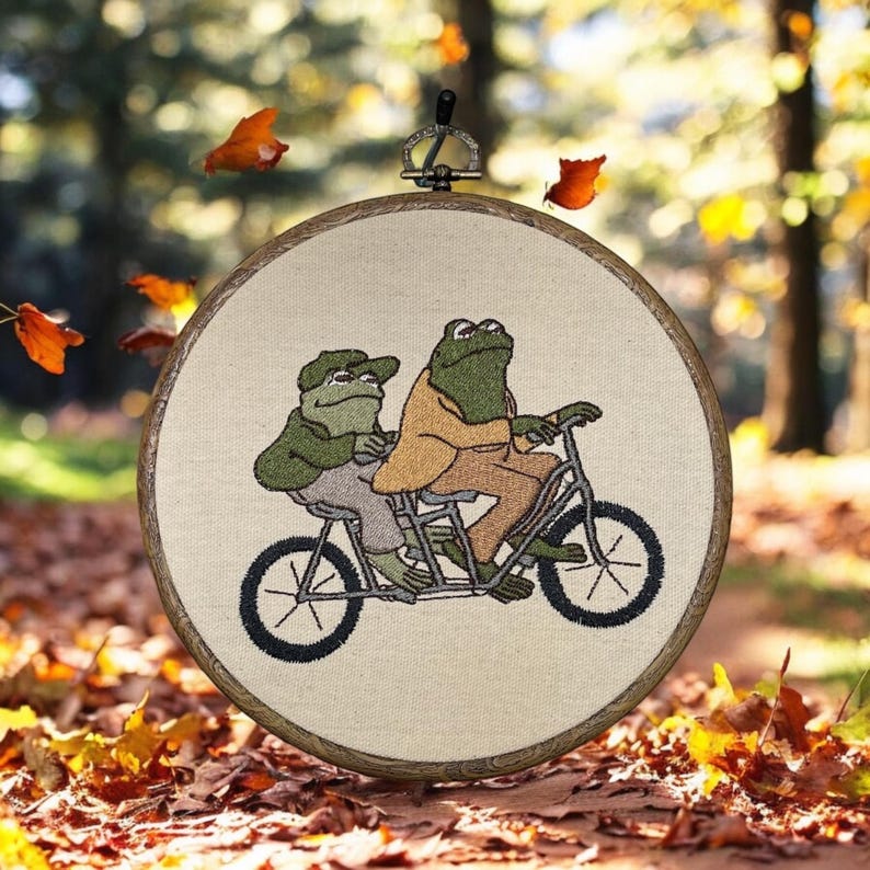 May include: Embroidered hoop art featuring two green frogs riding a bicycle. The frogs are wearing clothes and the bicycle has a basket.