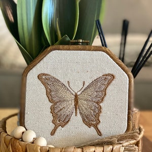 Puede incluir: Una mariposa marr&oacute;n bordada en un lienzo blanco en un marco de madera. El marco est&aacute; sentado en una cesta tejida con cuentas de madera.