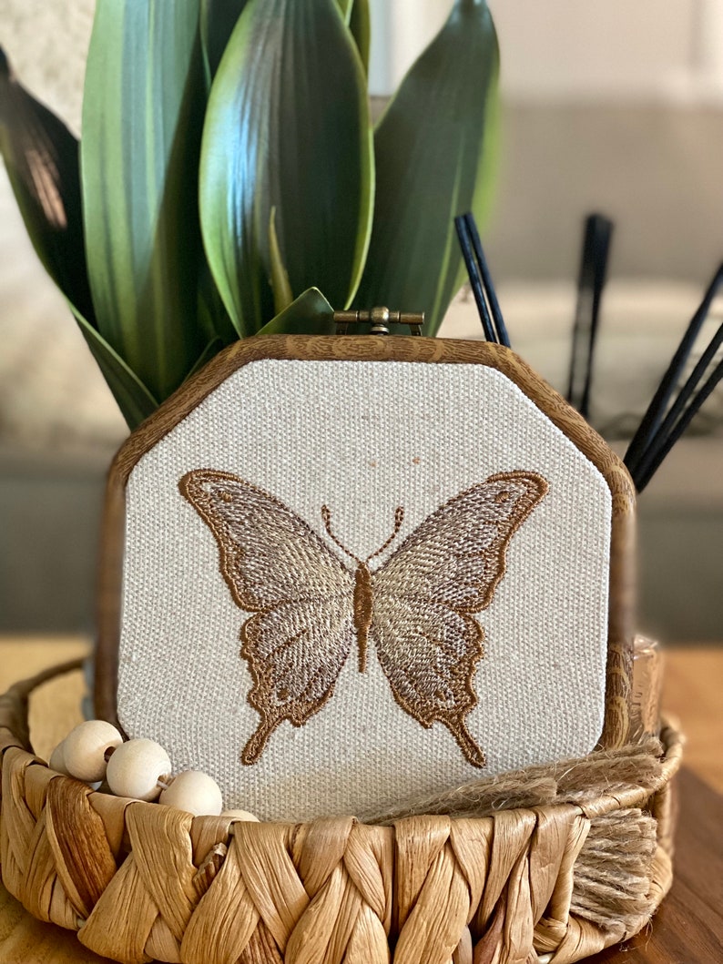 Puede incluir: Una mariposa marr&oacute;n bordada en un lienzo blanco en un marco de madera. El marco est&aacute; sentado en una cesta tejida con cuentas de madera.