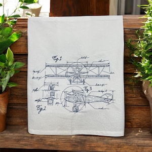 May include: A white kitchen towel with a blue embroidered design of a vintage airplane blueprint. The blueprint includes three different views of the airplane labeled "Fig 1", "Fig 2", and "Fig 3".