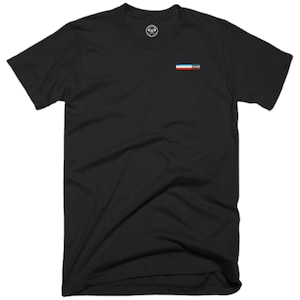 Könnte beinhalten: Schwarzes T-Shirt mit einer kleinen, bunten Flagge und dem Text "2003" auf der Vorderseite.