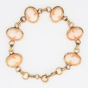 May include: A gold link bracelet featuring five oval-shaped cameos. Each cameo displays a carved profile of a person in cream-colored relief against a peach-toned background. The bracelet is secured with a circular spring ring clasp.