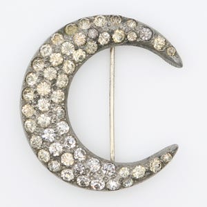 Peut inclure: Broche en forme de croissant de lune avec un cadre en métal argenté. La broche est incrustée de nombreux strass clairs et couleur champagne. Une épingle argentée est fixée au dos pour la fixation. La broche est probablement vintage ou antique.
