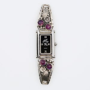 May include: A silver wristwatch with a rectangular black face. The watch has decorative floral and leaf designs with purple gemstones. The watch face displays the brand name "Hart" and Roman numerals.