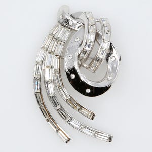 May include: A silver-toned metal brooch featuring a swirling, abstract design. The piece is adorned with numerous clear, rectangular baguette-cut crystals arranged in rows, accented by small, round, clear stones set into the polished metal frame.