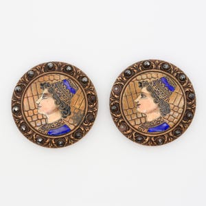 May include: Pair of round, gold-toned buttons with a raised, ornate border. Each button features a painted image of a woman's profile wearing a blue and gold hat. The buttons are decorated with black rhinestones.