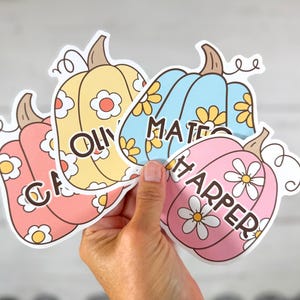 Puede incluir: Cuatro pegatinas con forma de calabaza coloridas con dise&ntilde;os florales y nombres: CA, Olly, Mateo y Harper. Las calabazas son en tonos rosa, amarillo y azul, con detalles de flores blancas y amarillas. Cada calabaza tiene un tallo marr&oacute;n y rizos.