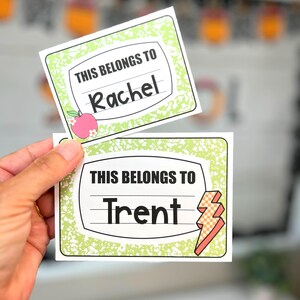 May include: Two rectangular name tags with a notebook paper design. The top tag reads "THIS BELONGS TO Rachel" with an apple graphic. The bottom tag reads "THIS BELONGS TO Trent" with a lightning bolt graphic. Both tags have a green border.