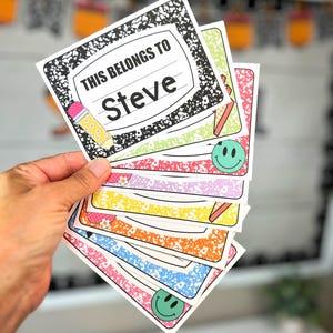 May include: A collection of colourful name tags with a notebook paper design. The top tag reads "THIS BELONGS TO Steve" with a pencil graphic. Other tags feature smiley faces and various patterns in pink, green, yellow, orange, and blue.