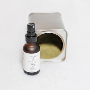 May include: A brown glass bottle with a black spray nozzle labeled "Refreshing Fresh Mist" sits next to a silver tin filled with green powder. The bottle contains 1 fl oz of product. The label has a minimalist design.