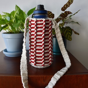 May include: A red and white striped crocheted bottle holder with a long beige strap. The bottle holder is on a wooden surface.
