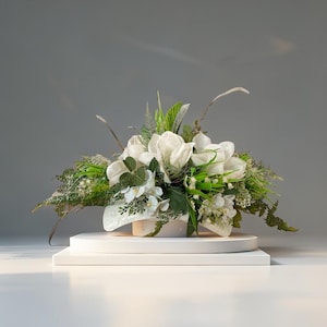 Può includere: Composizione floreale con fiori bianchi e fogliame verde, con accenti di steli secchi. L'arrangiamento è presentato su una piattaforma bianca, rotonda e a più livelli, su sfondo grigio. I fiori sono il fulcro.