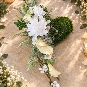 Könnte beinhalten: Ein herzförmiges Blumenarrangement mit einer Basis aus dichtem grünem Moos, verziert mit weißen Chrysanthemen, cremefarbenen Calla-Lilien und zarten weißen Blüten, ergänzt durch feines grünes Blattwerk und dünne Zweige.