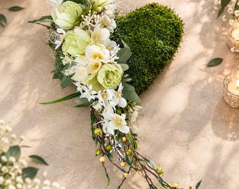 Puede incluir: Una decoración en forma de corazón cubierta de musgo verde, adornada con un grupo de flores artificiales de color crema y verde pálido, incluyendo peonías y pequeñas flores, con ramas marrones y bayas amarillas que sobresalen.