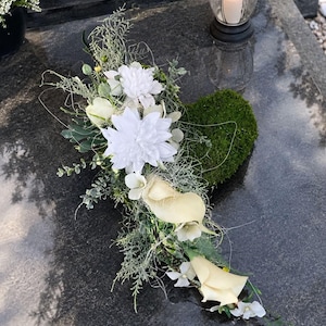 Könnte beinhalten: Ein herzförmiges Blumenarrangement mit leuchtend weißen Dahlien und cremefarbenen Calla-Lilien, akzentuiert durch grünes Moos, feines Blattwerk und zarte Zweige. Das Arrangement liegt auf einer dunklen, polierten Steinoberfläche.