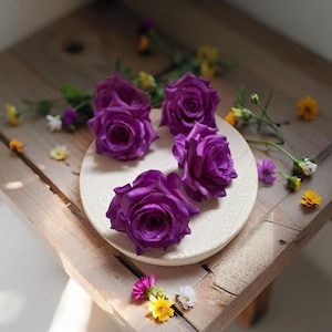 Könnte beinhalten: Vier lilafarbene Seidenrosen, die auf einem weißen Teller mit einem dekorativen Muster angeordnet sind. Der Teller liegt auf einer Holzoberfläche mit kleinen weißen und gelben Blumen, die um ihn herum verstreut sind.