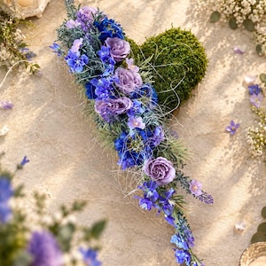 Könnte beinhalten: Ein herzförmiges Gesteck mit einer Basis aus dichtem grünem Moos, verziert mit einem kaskadierenden Arrangement aus blauen und violetten Blumen, darunter Rosen und Rittersporn, akzentuiert durch feine weiße Fasern und grünes Blattwerk.