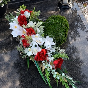 Könnte beinhalten: Ein herzförmiges Blumenarrangement mit leuchtend roten Rosen und weißen Freesien, akzentuiert durch grünes Moos und zartes Blattwerk. Das Arrangement ist auf einer dichten Moosbasis gestaltet und enthält lange, schmale grüne Blätter.