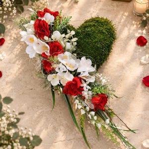 May include: A heart-shaped moss arrangement decorated with a cascading floral display of red roses, white freesia, and delicate greenery. The composition features a dense green moss base accented by vibrant red and white blossoms and foliage.
