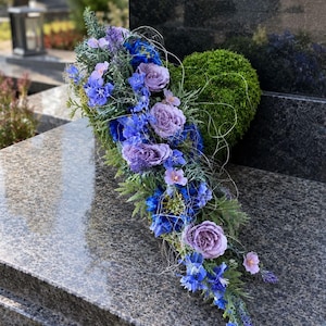 Könnte beinhalten: Ein herzförmiges Blumenarrangement mit einer Basis aus grünem Moos, verziert mit einer kaskadierenden Anordnung aus blauen und lavendelfarbenen Blumen, darunter Rosen und Rittersporn, akzentuiert durch feinen weißen Draht und grünes Laub.