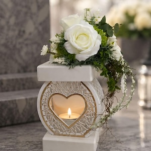 May include: A white, heart-shaped candle holder with gold trim and a lit candle inside. The holder is topped with white roses and greenery. The design is elegant and suitable for memorial or decorative purposes.