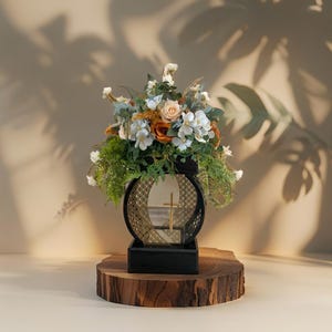 Puede incluir: Una urna negra con una cruz dorada y un arreglo floral de flores blancas, melocotón y naranja. La urna está sobre una base de madera.