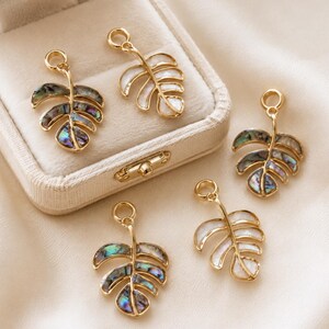 May include: A collection of five gold-toned metal leaf-shaped charms, each featuring a small circular loop at the top. Three charms are inlaid with iridescent abalone shell, while two charms are inlaid with smooth, white pearlescent material.