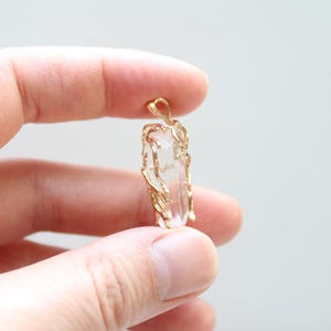 May include: A clear quartz crystal pendant with a gold-coloured wire wrap. The crystal is rectangular and suspended from a small loop. The gold wire wraps around the crystal, creating a decorative cage-like design. The pendant is held between fingers.