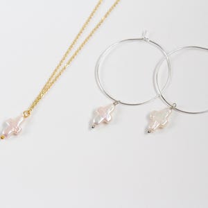 Puede incluir: Un conjunto de joyas que incluye un collar de cadena dorada y pendientes de aro de plata. Cada pieza tiene un colgante de cruz de perla.