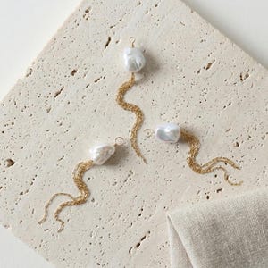 May include: Three pairs of earrings with a white, irregular-shaped pearl and a gold chain design. The earrings are displayed on a textured, light-colored surface, with a neutral-colored fabric in the corner.